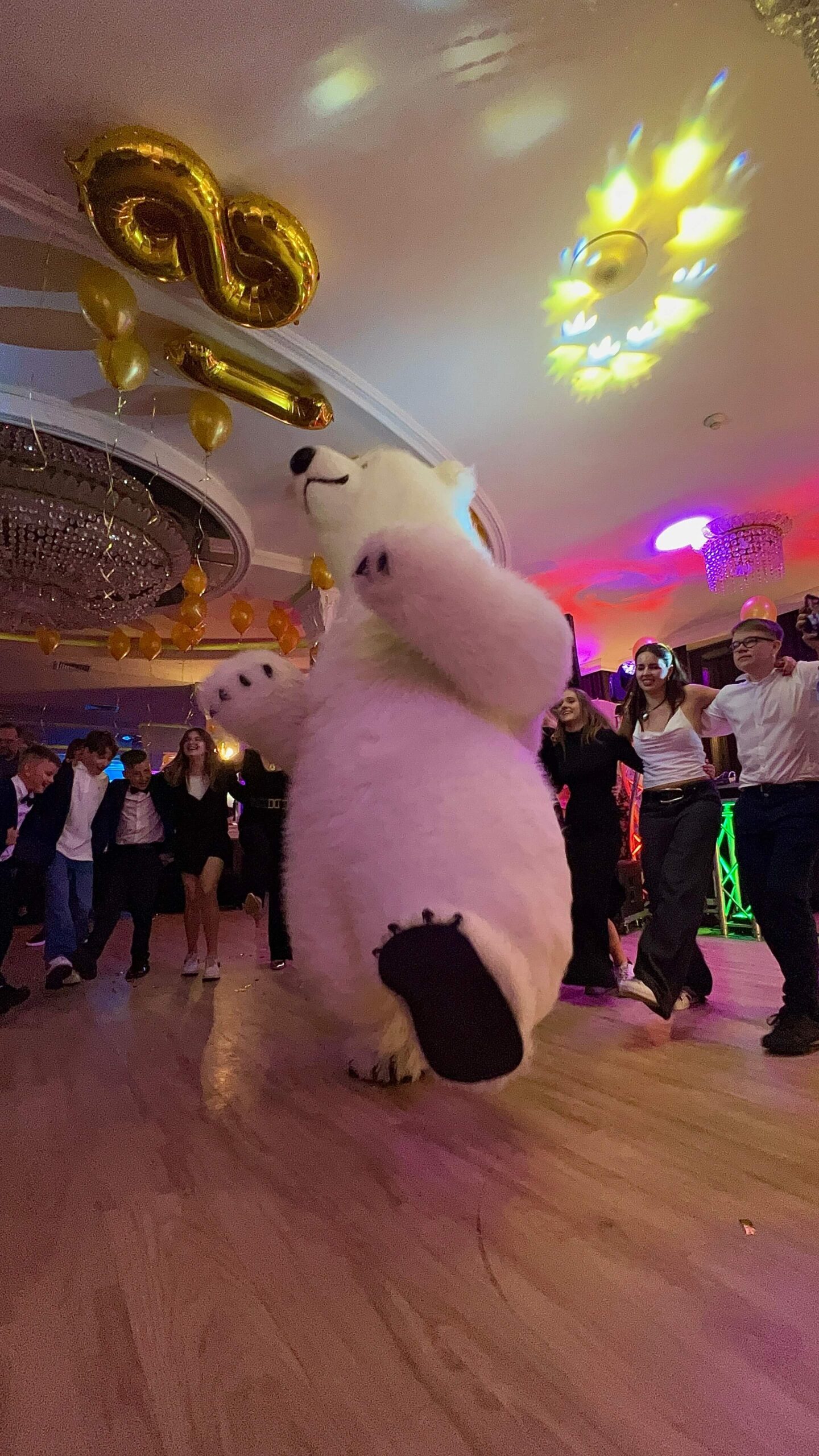 Person in a polar bear costume dancing at a party with golden '16' balloons and colorful lights in the background.