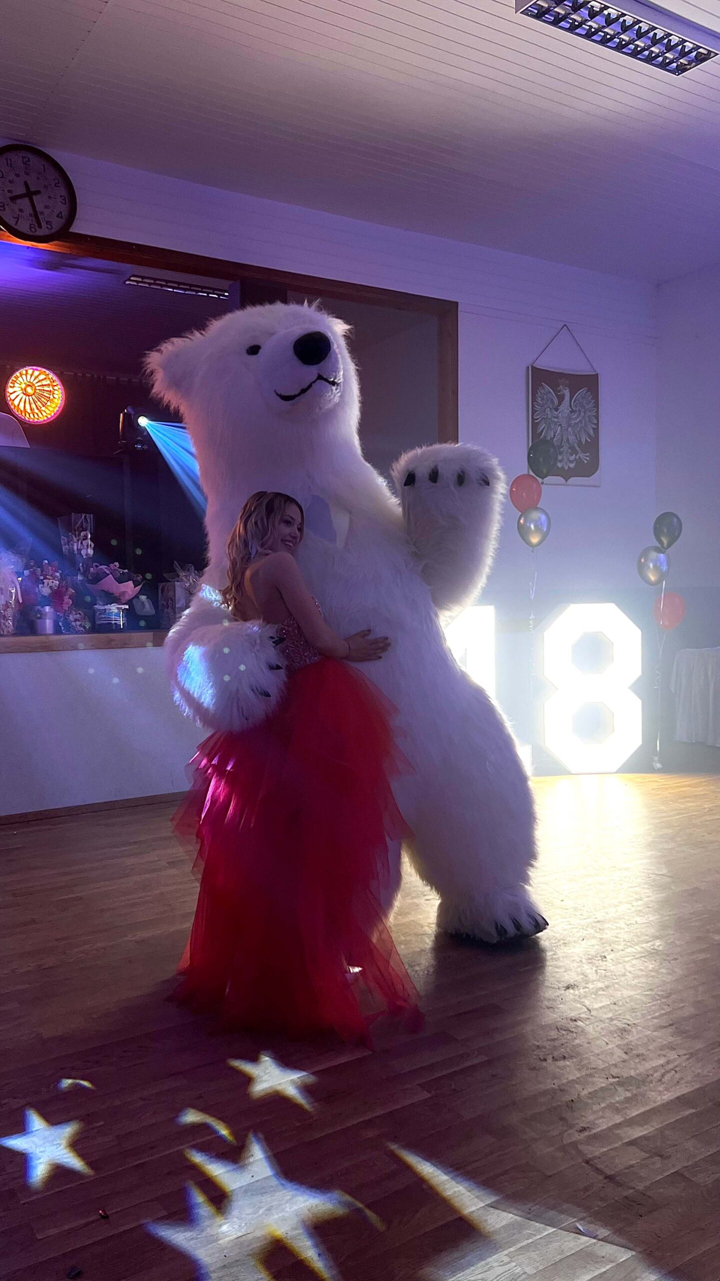 A young woman in a red dress embracing a person in a polar bear costume at a dance event, with '18' light up numbers and star-shaped lights on the floor.