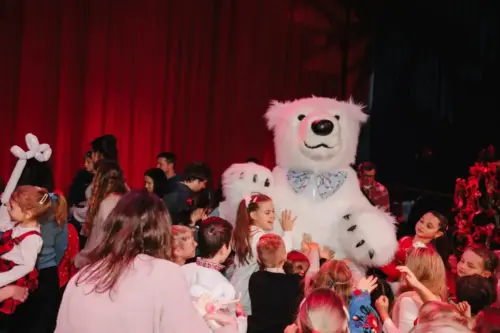A person in a polar bear costume seated on stage surrounded by a crowd of children and adults at a holiday gathering.