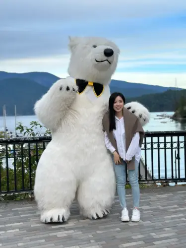  In this vibrant photograph, a man stands confidently next to a person in an oversized polar bear costume outside the entrance of the Metropolis at Metrotown shopping center. The bear, which is wearing a bright yellow bow tie, towers over the scene, creating a playful contrast with the urban environment. The shopping center's signage is prominently displayed in the background, with the modern architecture of the mall providing a contemporary backdrop. The clear sky and the mall's bustling atmosphere contribute to the lively setting of the image.