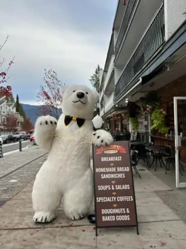 Person in polar bear costume waving beside a café menu sign on a sidewalk with mountains in the distance.