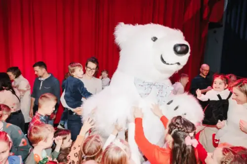 bear kids-AR078agQLJixPlq3Joyful children reaching out to a person in a polar bear mascot amidst an audience on a theater stage with red curtains.